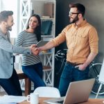 Good job! Confident young man standing near whiteboard and shaking hand to his colleague while young woman standing near them and smiling